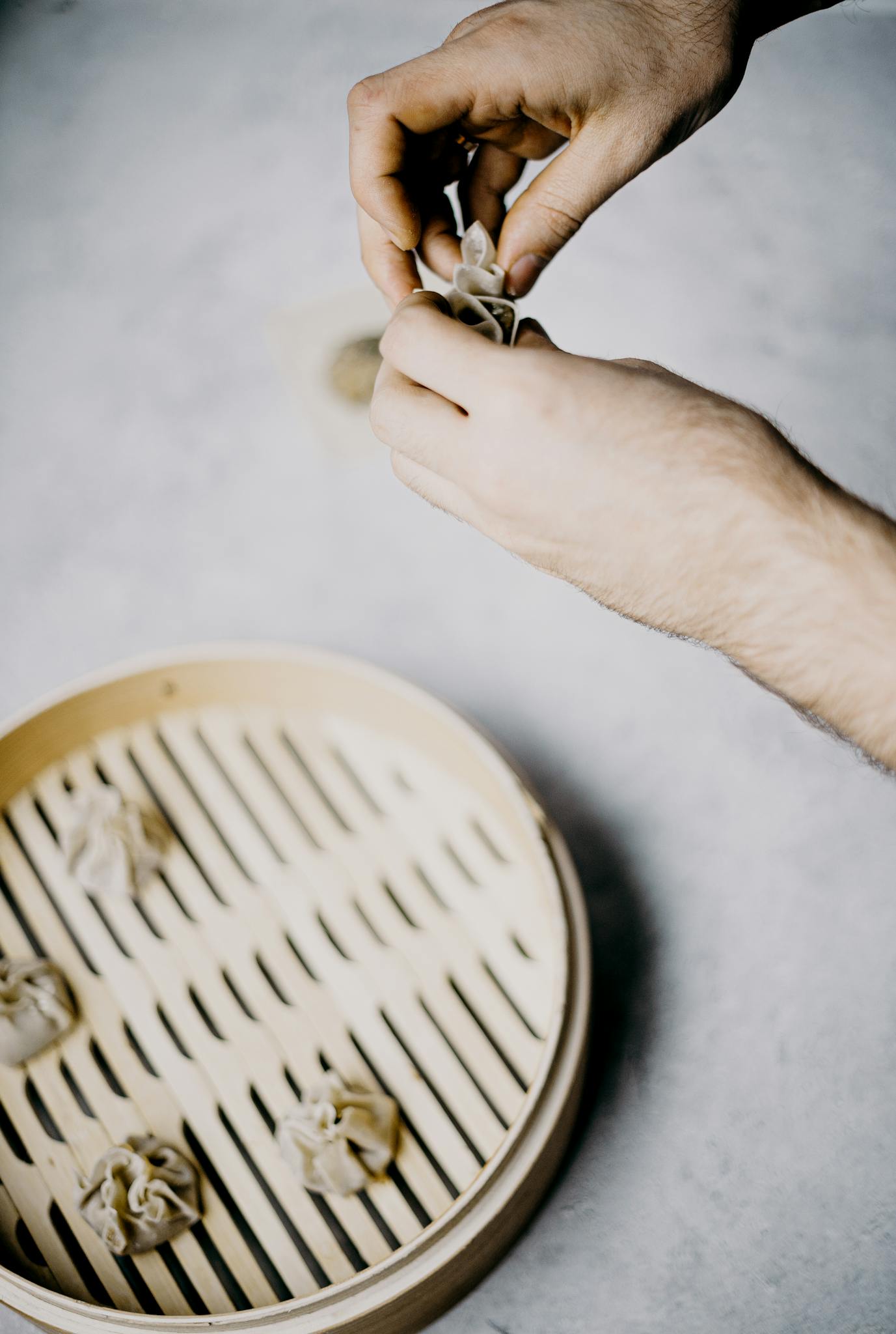 Close-up of hands making dumplings next to a bamboo steamer.